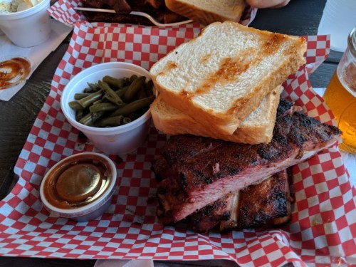 A plate with grilled ribs, toasted bread, green beans, and a small cup of sauce, served with a beer.