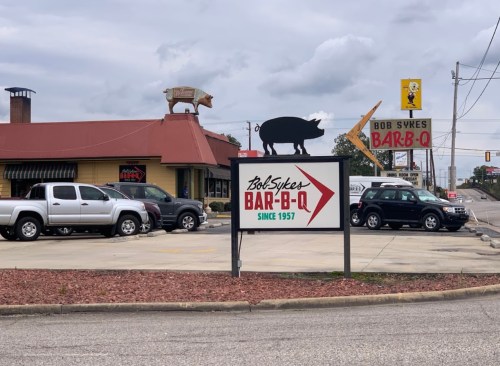 Sign for Bob Sykes Bar-B-Q with a pig statue on the roof, surrounded by parked cars and a cloudy sky.