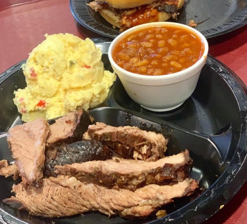 A black plate with sliced brisket, creamy potato salad, and a cup of baked beans.