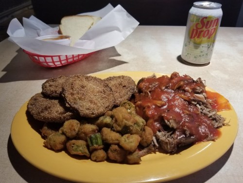 A plate of fried green tomatoes, pulled pork with barbecue sauce, fried okra, a slice of bread, and a can of Sun Drop.