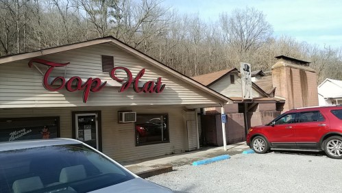 Exterior of the Top Hat building with a sign, surrounded by trees and parked cars in a gravel lot.