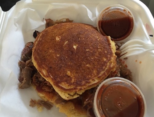A plate with pulled pork topped with a cornbread pancake and two cups of barbecue sauce on the side.