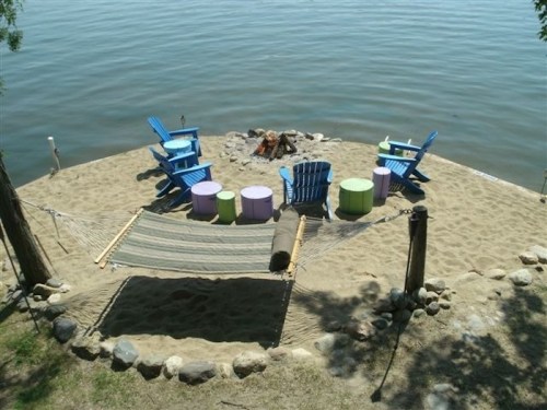 A sandy lakeside area with colorful chairs, a hammock, and a fire pit surrounded by water and trees.
