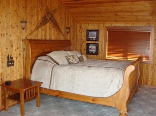Cozy wooden bedroom featuring a sleigh bed, wooden walls, and framed pictures. Natural light from a window.