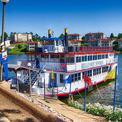 A colorful riverboat named "Belle" docked by the shore, surrounded by greenery and buildings under a clear blue sky.