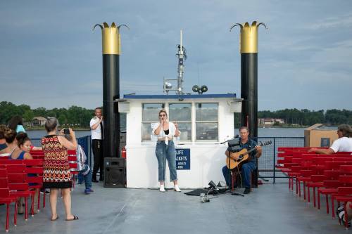 A woman sings on a boat while a guitarist plays; passengers enjoy the performance from red chairs.