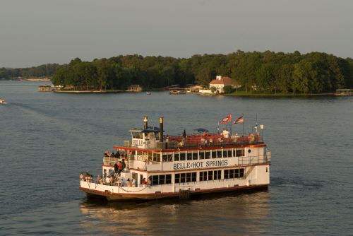 A riverboat named "Belle of Hot Springs" cruises on a calm lake, surrounded by green trees and a distant shoreline.