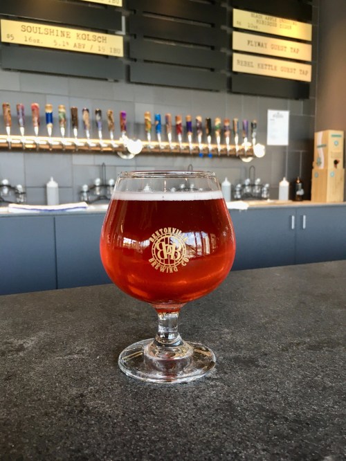 A glass of amber beer sits on a dark countertop, with a brewery tap wall in the background.