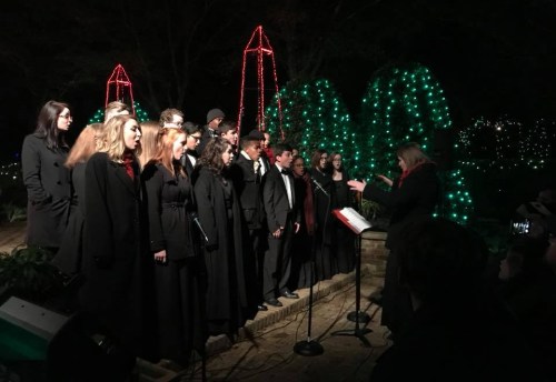 A choir in black attire performs outdoors at night, surrounded by festive green and red lights.