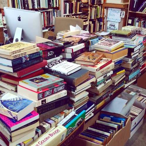 A cluttered table stacked high with various books in a cozy bookstore, with a computer in the background.