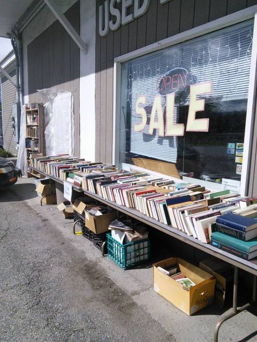A table outside a used bookstore displays various books for sale, with boxes underneath and an "Open Sale" sign.