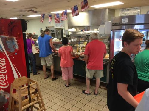 A busy diner with customers lining up at the counter, flags hanging from the ceiling, and a soda machine in the foreground.