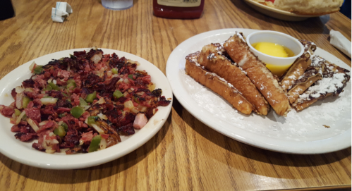 A plate of hash browns with diced meats and vegetables next to a plate of French toast with syrup and powdered sugar.
