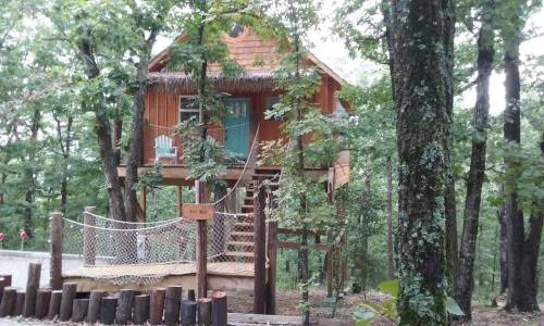 A wooden treehouse nestled among trees, featuring a porch, stairs, and a sign labeled "The Nest."