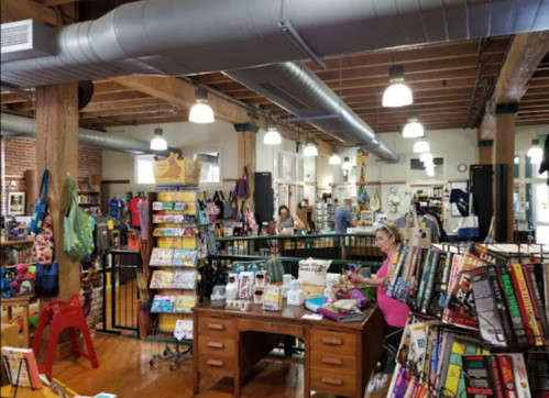 A cozy bookstore with wooden beams, shelves of books, and a woman at a desk surrounded by colorful merchandise.