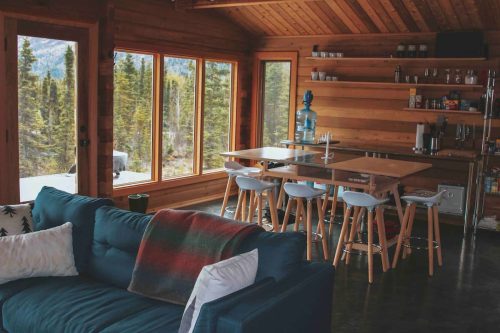 Cozy wooden cabin interior with a blue sofa, dining table, and large windows showcasing a forest view.