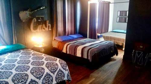 A cozy bedroom with three beds, dark curtains, and a guitar decor piece on the wall. Warm lighting adds ambiance.