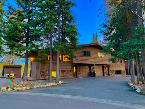 A two-story brown house surrounded by tall trees, with a stone foundation and a paved driveway at dusk.