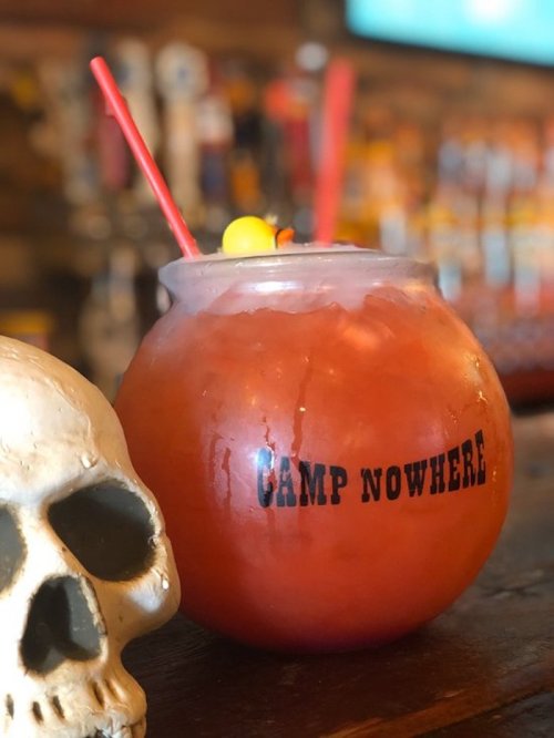A colorful cocktail in a fishbowl glass with a red straw and cherry, next to a decorative skull.
