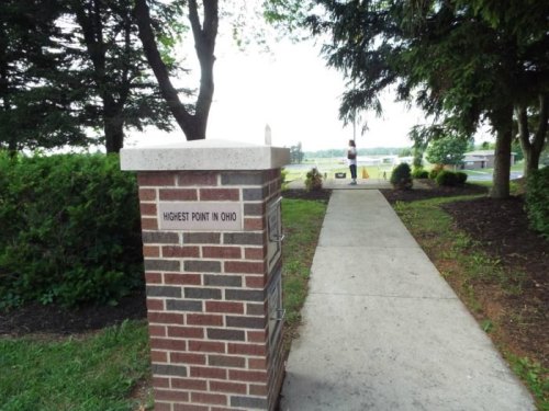 A brick monument labeled "Highest Point in Ohio" beside a paved path leading to a grassy area and trees.