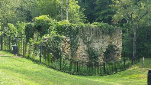 Ruins of a stone wall covered in greenery, surrounded by a black fence and lush grass in a wooded area.