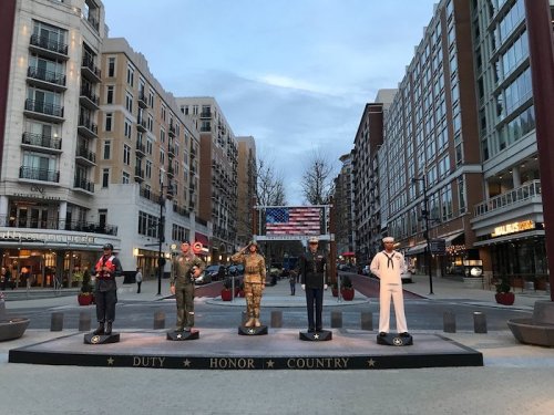 Statues of military personnel in uniform stand in a plaza, with an American flag and buildings in the background.