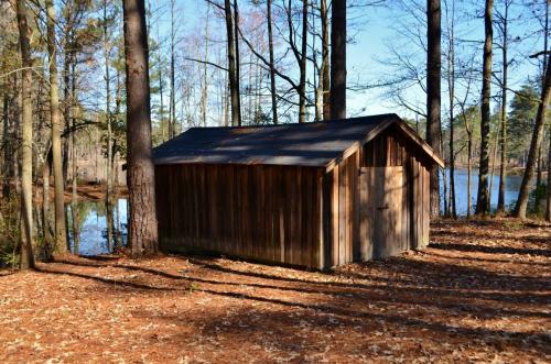 A wooden shed beside a lake, surrounded by trees and fallen leaves in a serene outdoor setting.