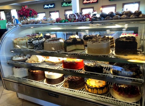 A display case filled with various colorful cakes and desserts in a bakery, decorated for the holidays.