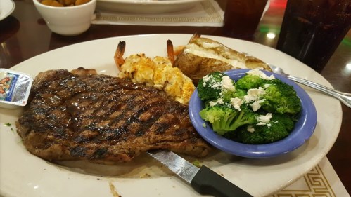 A plate featuring a grilled steak, shrimp, a baked potato, and a side of broccoli with feta cheese.