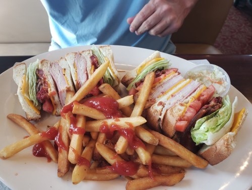 A plate with two sandwiches stacked with meats and veggies, accompanied by a pile of fries and a small cup of coleslaw.