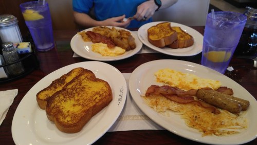 A table with two plates of breakfast: French toast, bacon, eggs, sausage, and hash browns, with drinks in the background.