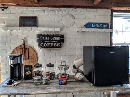 A cozy kitchen scene featuring a coffee maker, jars, and a mini fridge against a white brick wall with decorative signs.