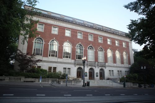 A large brick library building with arched windows, surrounded by trees and a sidewalk in the foreground.