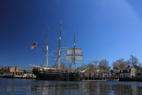 A tall ship with sails raised docked by a waterfront, surrounded by trees and buildings under a clear blue sky.