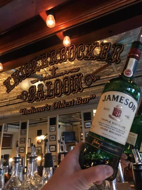 A hand holds a bottle of Jameson whiskey in front of the Knickerbocker Saloon sign, "Indiana's Oldest Bar."