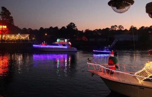 Colorful boats decorated with lights glide through a calm waterway at dusk, creating a festive holiday atmosphere.
