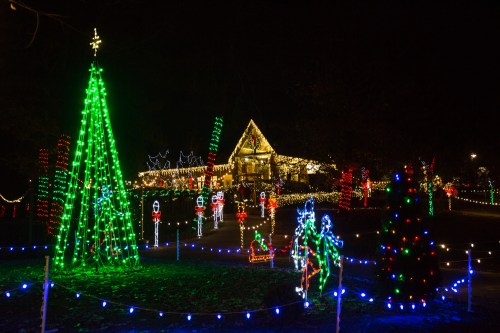 Colorful Christmas lights illuminate a festive scene with decorated trees and a cozy house in the background.