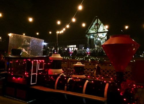 A festive train decorated with lights, set against a backdrop of a house adorned with holiday decorations at night.