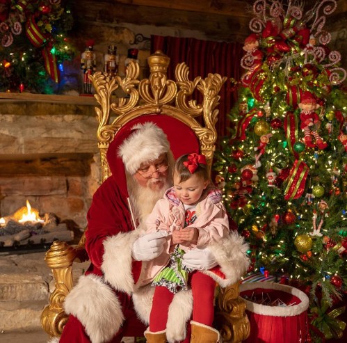Santa Claus sits in a golden chair with a young girl, surrounded by festive decorations and a Christmas tree.