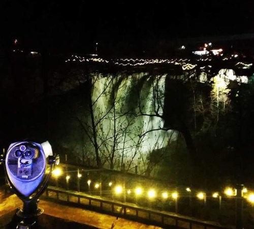 Night view of a waterfall illuminated by lights, with a viewing telescope in the foreground.