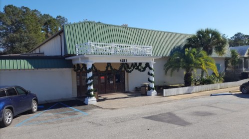 A building with a green roof, decorated with garlands, featuring palm trees and a parking area in front.