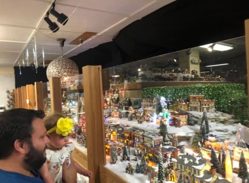 A man and a child admire a festive miniature village display with lights and snow.