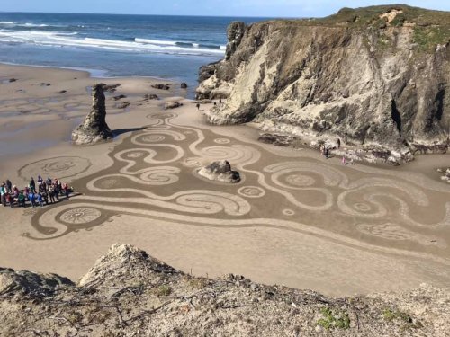 Aerial view of intricate sand art on a beach, with people gathered around a large rock formation. Ocean in the background.