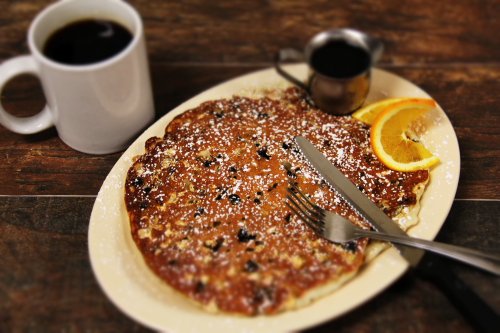 A plate of pancakes topped with powdered sugar, served with orange slices and syrup, alongside a cup of coffee.