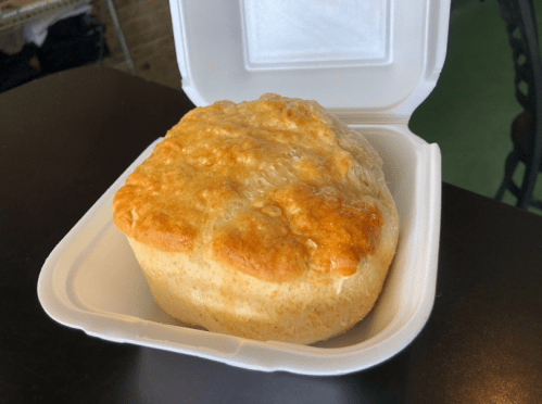 A golden, flaky biscuit in a white takeout container, resting on a dark table.