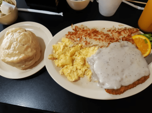 A plate of scrambled eggs, hash browns, chicken fried steak with gravy, and a biscuit, garnished with an orange slice.