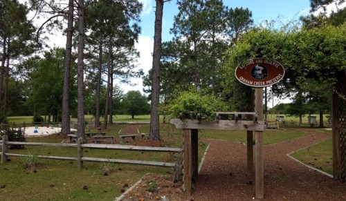 A park entrance with a wooden sign, surrounded by trees and a pathway leading to picnic areas and open green space.