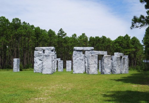 A replica of Stonehenge made of gray stones, set in a grassy area surrounded by tall trees under a blue sky.