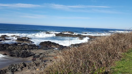 A scenic view of rocky coastline with waves crashing, under a clear blue sky and distant sailboat on the horizon.