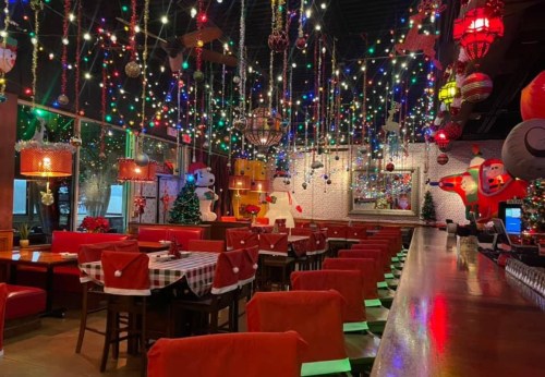A festive restaurant decorated with colorful lights, snowmen, and Santa figures, featuring red tablecloths and a long bar.